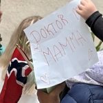 Barn holder opp en hjemmelaget plakat med teksten «Doktor mamma» og små tegninger av hjerter, ved siden av et norsk flagg. Fotokreditering kan stå separat: Foto: Øyvind Byrkjedal-Bendiksen