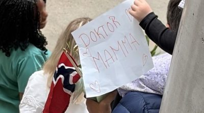 Barn holder opp en hjemmelaget plakat med teksten «Doktor mamma» og små tegninger av hjerter, ved siden av et norsk flagg. Fotokreditering kan stå separat: Foto: Øyvind Byrkjedal-Bendiksen