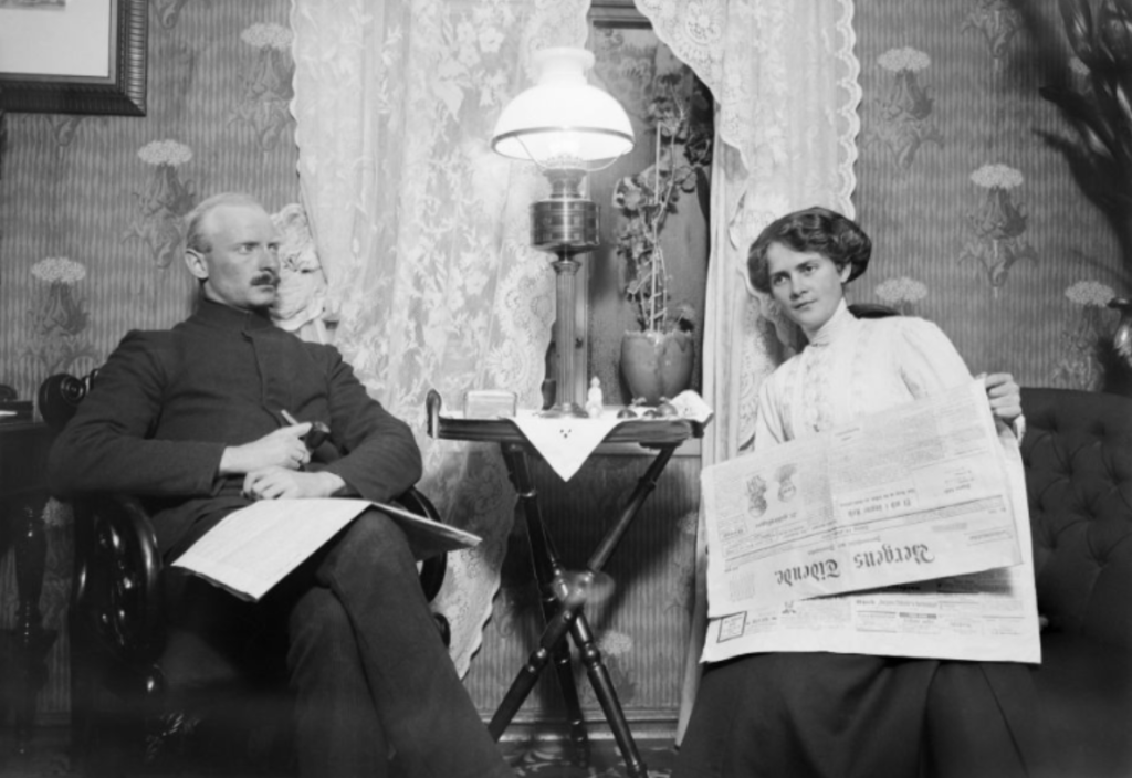 Alternativ tekst: Svart-hvitt fotografi av et interiør fra tidlig 1900-tall. To personer sitter på hver sin side av et lite bord med en lampe. Personen til venstre sitter i en lenestol med papir eller notatblokk i fanget, mens personen til høyre holder opp en avis. Rommet har mønstret tapet, blondegardiner og dekorative møbler.